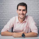 Confident young man in a pink shirt smiling at the camera against a brick wall backdrop.
